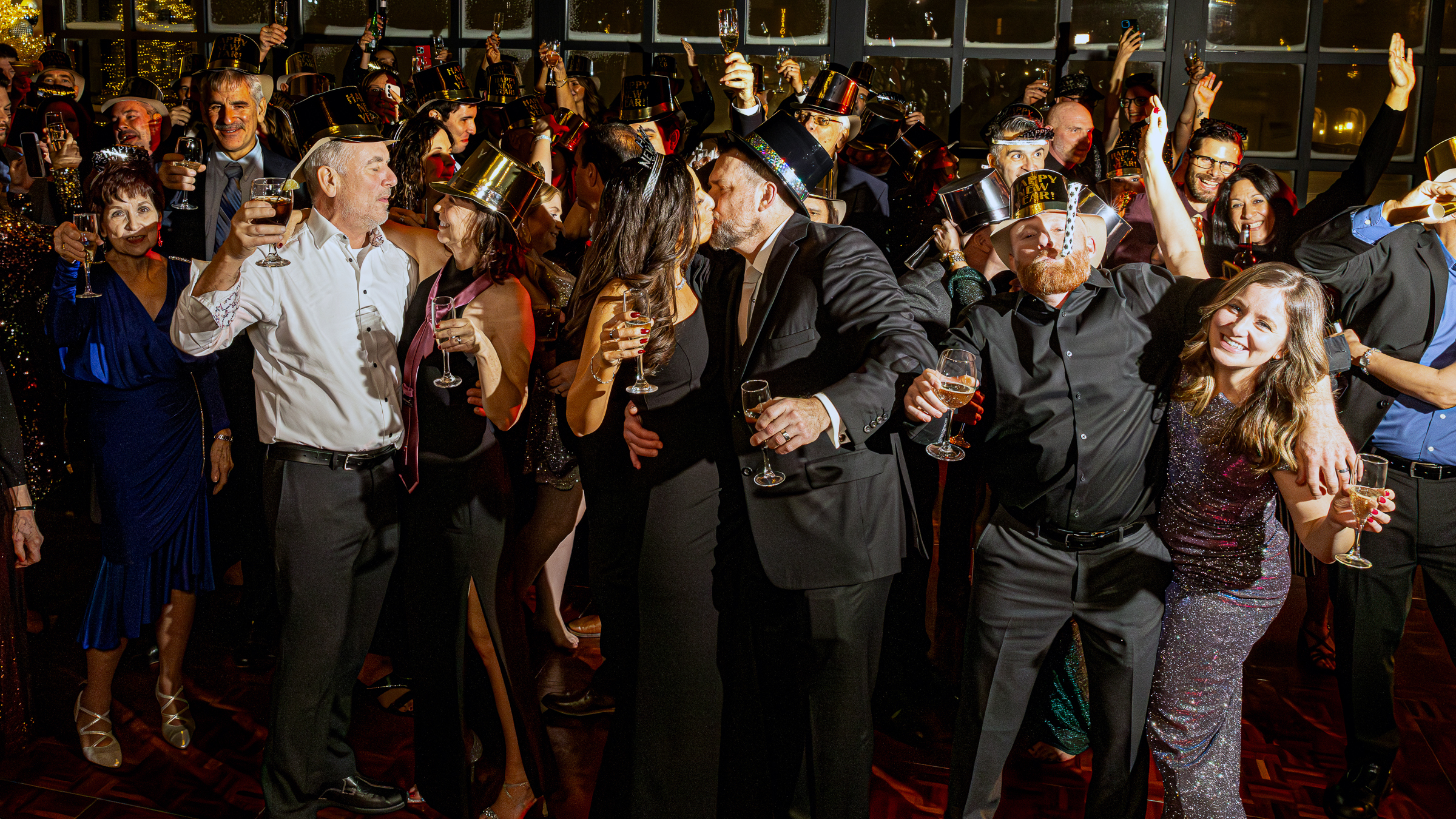 Crowd of people celebrating New Year's Eve with champagne toasts, party hats, and a couple kissing at midnight.