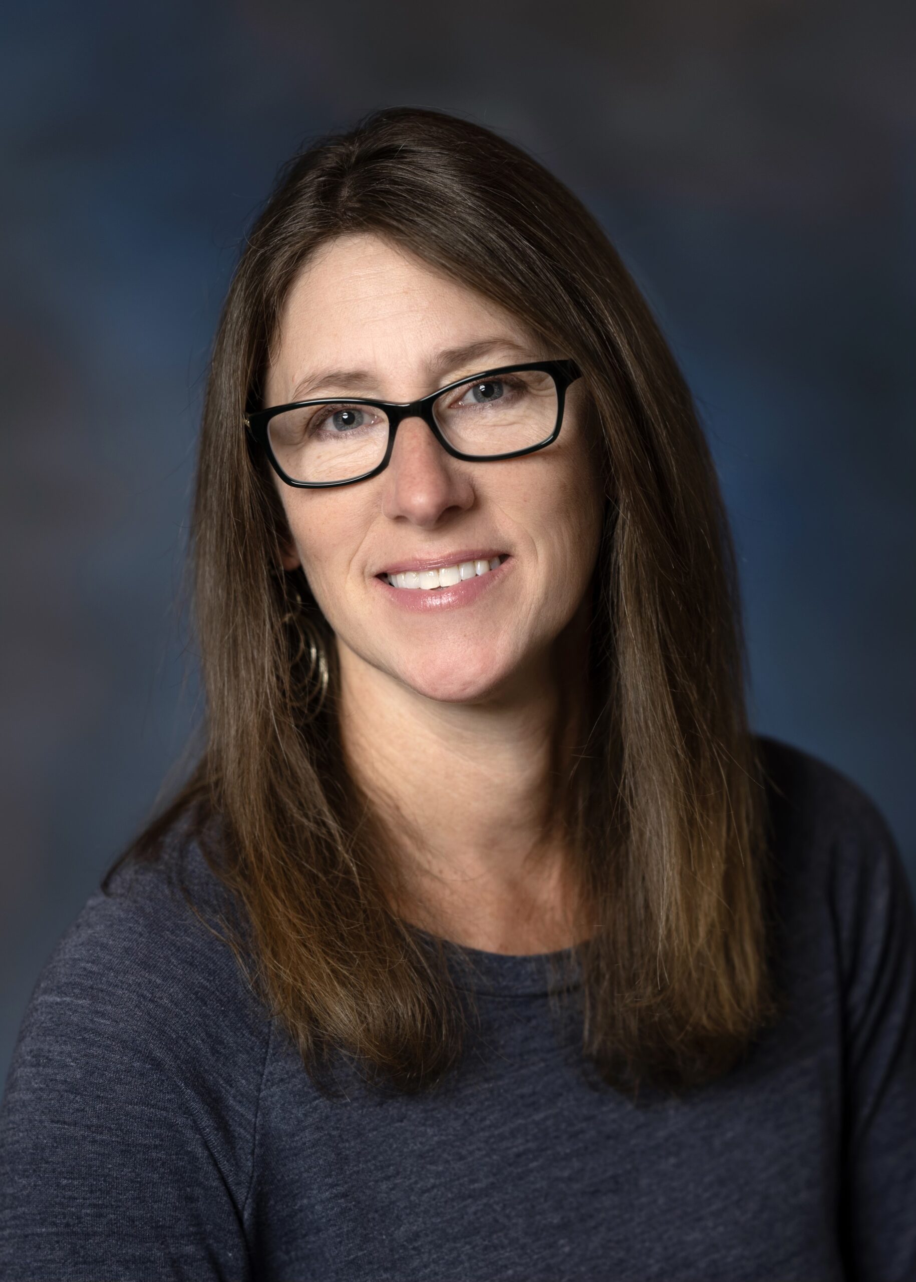 Andrea Bartel, a professional photographer with long brown hair and glasses, smiling in a portrait against a blue mottled background.