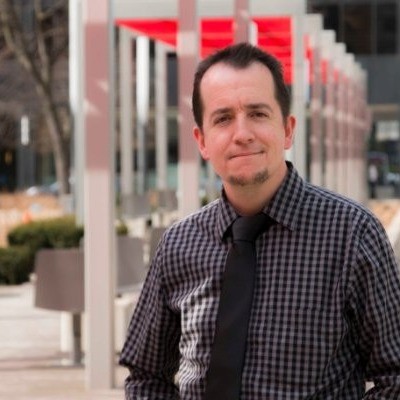 Chris Stabile, a professional cinematographer, wearing a checkered shirt and tie, posing outdoors in an urban setting with red architectural accents.