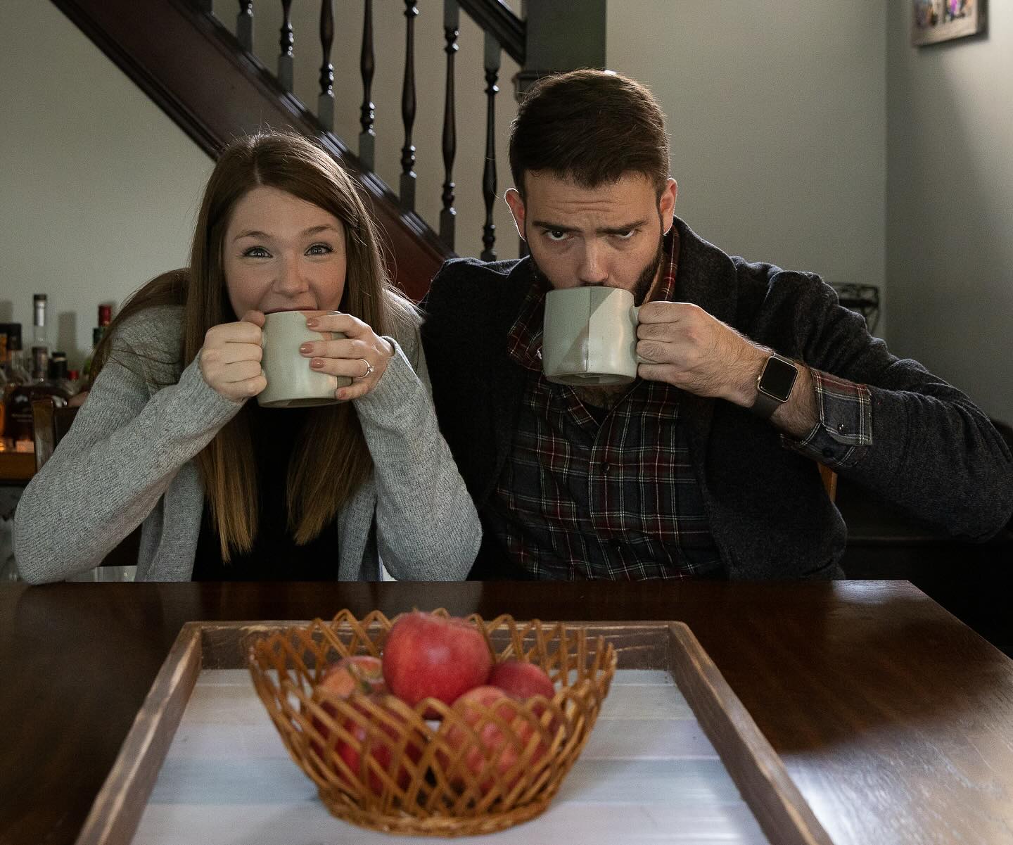 Erin Stewart Burke and husband Mason sitting at a wooden table, playfully peeking over the rims of light green ceramic mugs with a basket of red apples in the foreground.