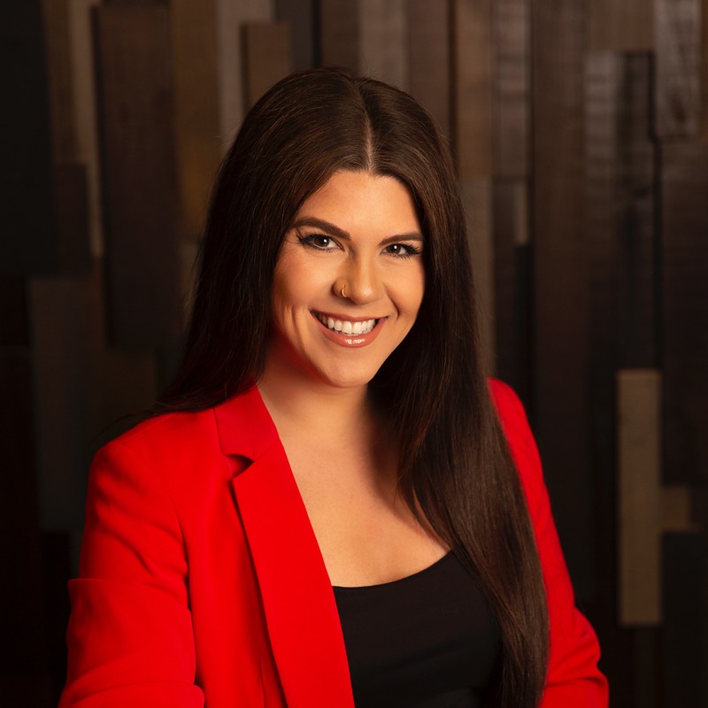 Katie Cole smiling, wearing a vibrant red blazer over a black top, with long dark hair and a nose ring.