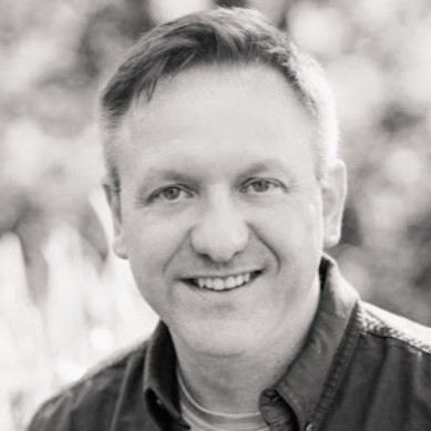 Black and white headshot of a smiling Paul Perhacs with a soft-focus natural background.