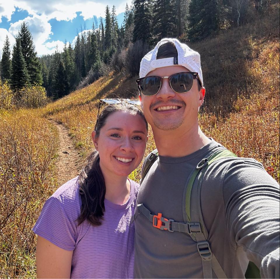 A smiling couple taking a selfie while hiking a mountain trail surrounded by yellow autumn grass and evergreen trees under a blue sky.