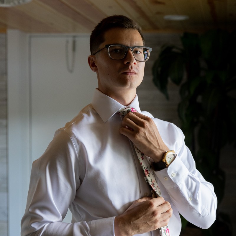 Zach Tancak in a white dress shirt and floral tie, adjusting his collar while wearing glasses and a watch.