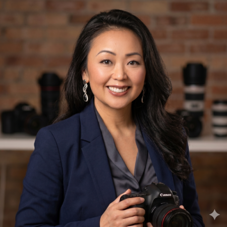 Professional headshot of Lana Perhacs, a female photographer with long dark hair, wearing a navy blazer and smiling while holding a Canon DSLR camera against a blurred studio background.
