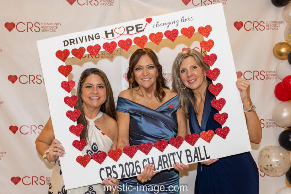 Three smiling women pose at a charity event behind a photo frame decorated with red hearts, reading 'DRIVING HOPE' and 'CRS 2026 CHARITY BALL'.